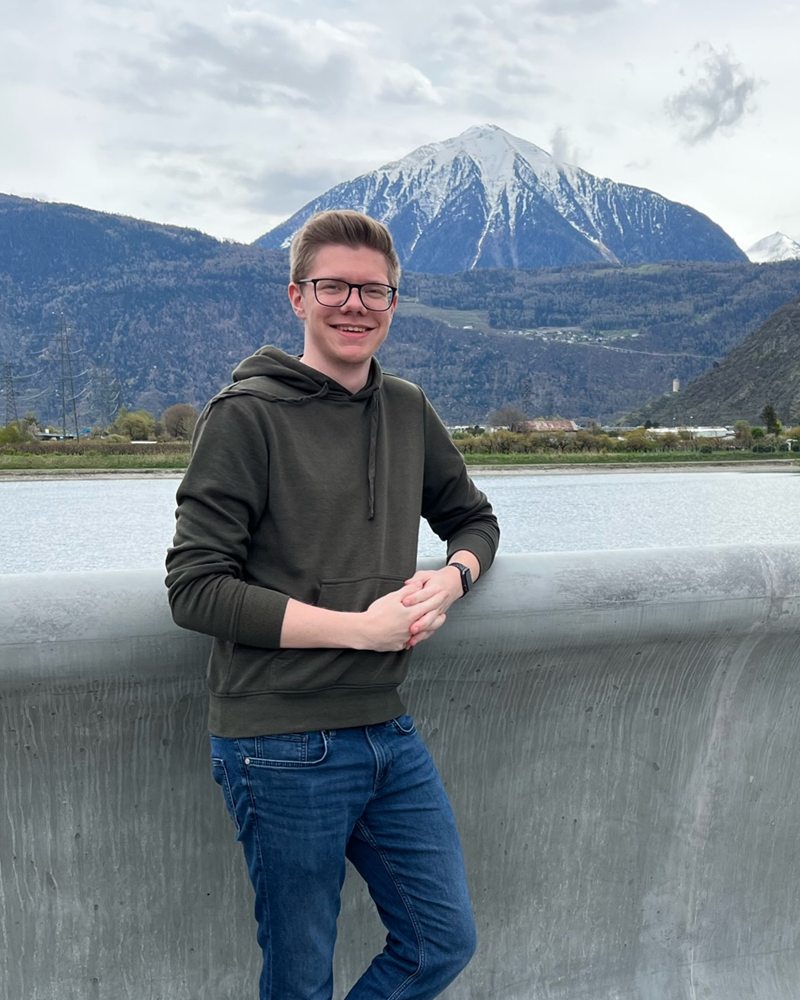 Maximilian Imsiecke lehnt lächelnd an einer Mauer, dahinter ein See und schneebedeckte Berge
